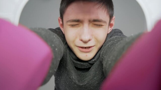 Teenager in rubber gloves grimaces with disgust as he plunges hands into toilet to unclog blocked pipe, wrinkling his nose at foul odor. Boy cleans toilet. View from toilet bottom. Cleanliness and