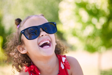 Girl, child and laughing with sunglasses in park, garden or nature to relax on holiday in summer. Kid, happy and excited with thinking, joke and comic memory in sunshine with funny ideas on vacation
