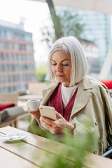 Attractive older woman checking smartphone, drinking coffee at coffee shop. Mature woman spending free time outdoors, in cafe, waiting for girl friends.