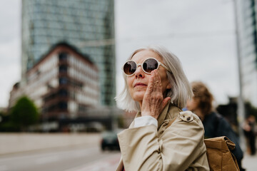 Portrait of stylish mature woman with gray hair on city street. Older woman in sunglasses waiting for public transport.