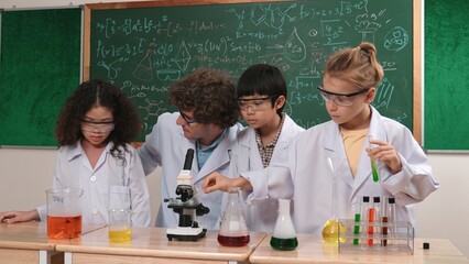 Smart caucasian teacher looking under microscope while diverse children doing experiment at STEM...
