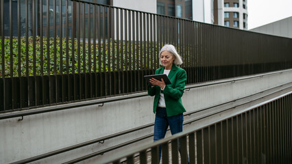 Mature businesswoman scrolling on tablet. Beautiful older woman with gray hair walking down city street.
