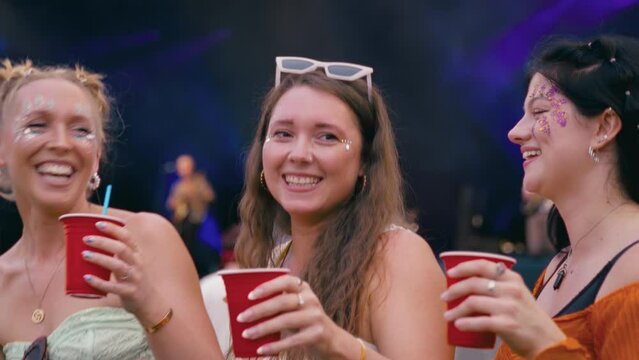 Three female friends wearing glitter holding drinks having fun dancing at outdoor summer music festival - shot in slow motion 