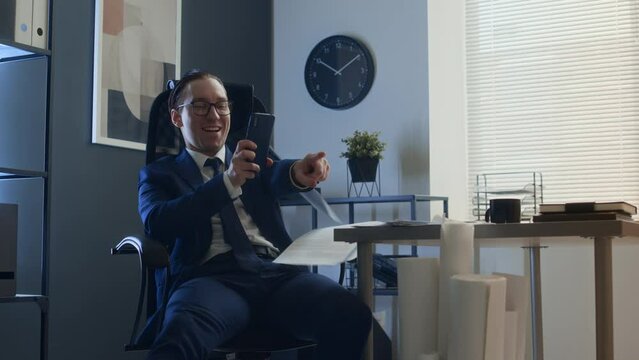 Medium shot of mean career guy in business suit, tie and glasses sitting in chair at desk, using smartphone, then tripping up colleague walking by with stack of papers and laughing viciouslyMedium sho