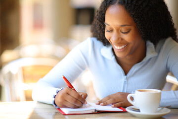Happy black woman writing in agenda in a coffee shop terrace