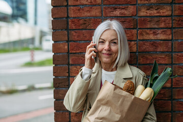 Mature woman making phone call, going home from supermaket with gorceries. Beautiful older woman with gray hair standing on city street.