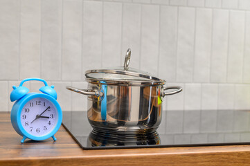 Czas gotowania, zegar stoi na blacie kuchennym obok stalowego garnka z gotującym się obiadem  - obrazy, fototapety, plakaty