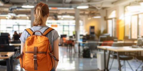 Rear view of a young female student with a bright orange backpack looking at a spacious coworking space.