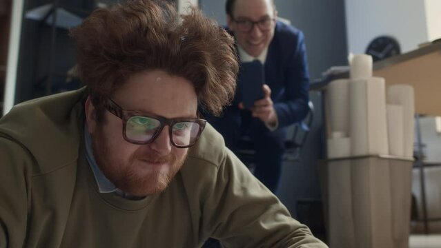 Medium close-up shot of red-haired nerdy male office worker raising head from floor after getting tripped, and bullying male colleague laughing and filming him on smartphoneMedium close-up shot of red
