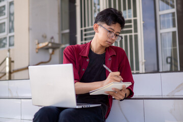 Asian college student studying on laptop at campus.  Man writing on a note book and working on laptop. Educational concept