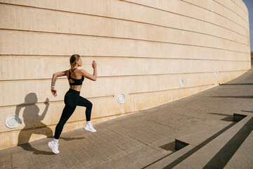 Fototapeta na wymiar Side view of woman in sportswear is running on buildings background. Active lifestyle concept