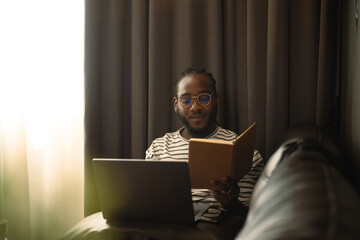 Shot of African man freelancer reading notes while working with laptop on sofa