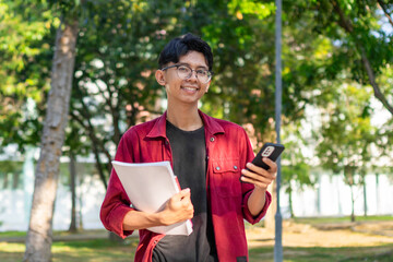 Young Asian college student using smartphone with happy expression. A male smiling while holding his phone and books at the public park. copy space