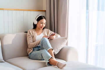 Happy young Asian woman with mobile phone listening music in headphones and relaxing on safa at home