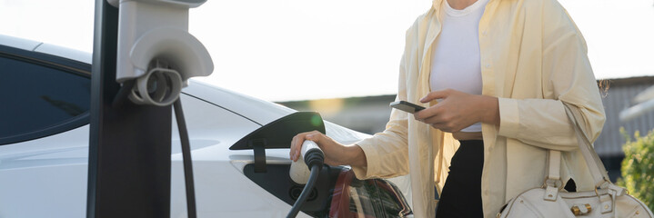 Woman holding shopping bag and use smartphone to pay for electricity to recharge EV car battery...