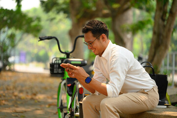 Happy young businessman sitting on bench near his bicycle using mobile phone