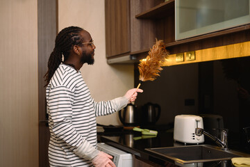 Young African man wiping dust on cabinet with a feather duster. Housekeeping concept