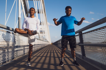 Two african-american friends are exercising on the bridge in the city. They are warming up for jogging. - 791405929