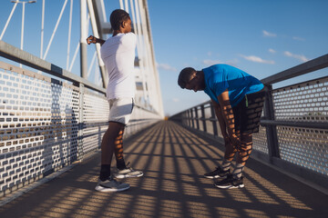 Two african-american friends are exercising on the bridge in the city. They are warming up for jogging. - 791405945