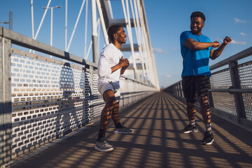 Two african-american friends are exercising on the bridge in the city. They are warming up for jogging. - 791405957