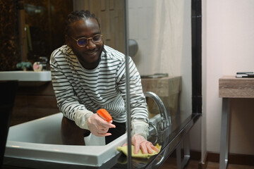 30s African guy in rubber gloves washing bathtub and faucet in a modern bathroom