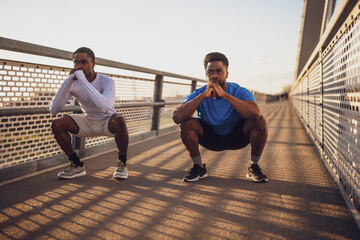 Two african-american friends are exercising on the bridge in the city. They are doing squats. - 791406371