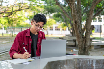 Asian college student studying on laptop at campus outdoor park.  Man writing on a note book and working on laptop. Educational concept