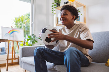 Happy boy holding soccer ball watching match at home