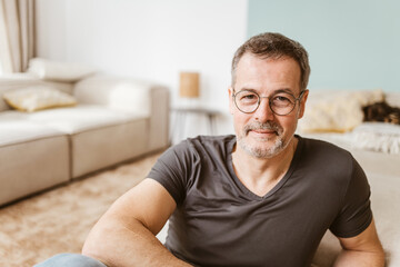 Smiling senior man with glasses and t-shirt sitting on the living room floor - 791411775