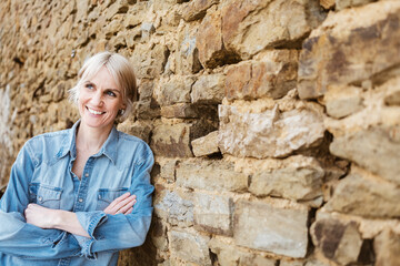Confident blonde woman in denim standing by a stone wall, smiling joyfully - 791413171