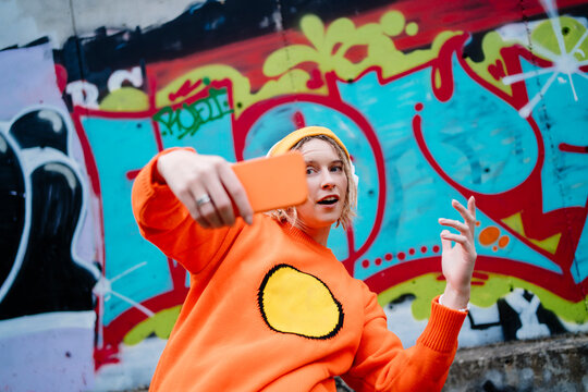 Young woman gesturing and taking selfie through smart phone in front of graffiti wall