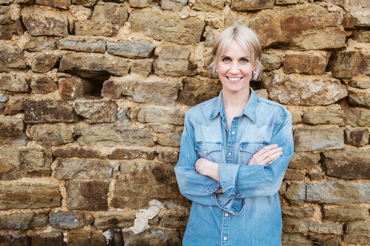 Confident blonde woman with short hair and denim shirt, smiling outdoors with crossed arms in front of a stone wall