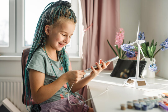 Smiling girl plugging smart phone near wind turbine model at table