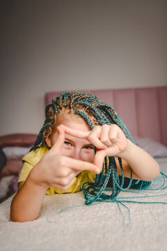 Happy girl with turquoise dyed braided hair lying on bed making finger frame at home