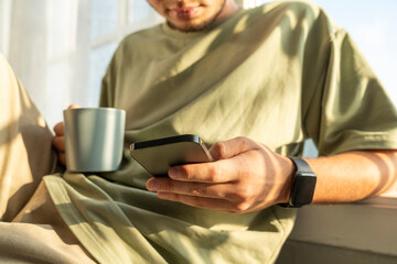 Young man holding coffee cup and using smart phone at home