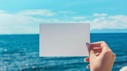 A woman's hand holds a white blank piece of paper against the background of the sea. Mockup, product advertisement, invitation flyer