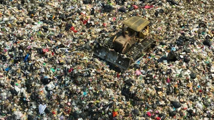 Fotobehang From above, a sprawling landfill sprawls, dotted with heaps of assorted refuse. A lone yellow bulldozer toils amidst the sea of waste, a testament to ongoing efforts in waste management.  © Punyawee