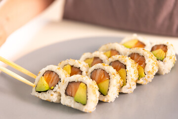 A plate of sushi with a person's hand in the background. The sushi is made with avocado and salmon