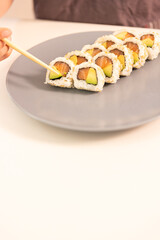 A plate of sushi with a person holding a chopstick. The plate has a variety of sushi rolls, including some with avocado. Concept of enjoyment and indulgence, as the person is about to eat the sushi