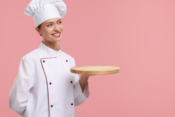 Happy woman chef in uniform holding empty wooden board on pink background, space for text