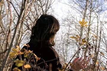 Young woman with dark hair admiring yellow autumn leaf in front of leafless bushes - 791434702