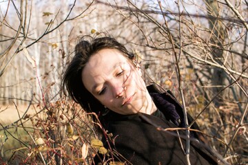 Contemplative woman in black coat surrounded by bushes, lost in thought, face peeking through branches - 791434798
