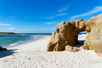 Arche naturelle sculptée dans la roche, encadrant une plage de sable blanc et des eaux turquoises, sous un ciel bleu, en Finistère Nord, Bretagne. - obrazy, fototapety, plakaty