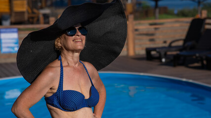 An elderly woman in a huge straw hat and sunglasses is sunbathing by the pool. Retiree on vacation.