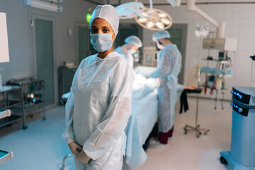 Portrait of skilled African-American female doctor standing posing looking at camera in light operating theater. Diverse team of surgeons performing operation on background. Concept of emergency. - Powered by Adobe