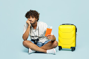 Full body sad traveler Indian man wear white casual clothes sit hold bag passport ticket isolated on plain blue background. Tourist travel abroad in free time rest getaway. Air flight journey concept.