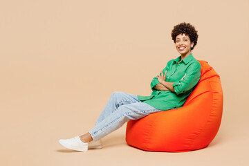 Full body young woman of African American ethnicity wear green shirt casual clothes sit in bag chair hold hands crossed folded look camera isolated on plain pastel beige background. Lifestyle concept.