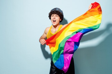 Young fun happy gay Latin man wear mesh tank top hat clothes hold striped rainbow flag sing isolated on plain pastel light blue cyan background studio portrait. Pride day June month love LGBT concept. - obrazy, fototapety, plakaty