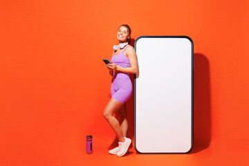 Full body young fitness trainer woman sportsman wears top shorts purple clothes in home gym big blank screen use mobile cell phone isolated on plain orange background. Workout sport fit abs concept.