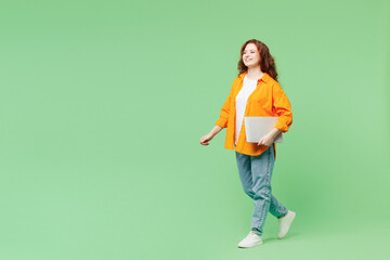 Fototapeta na wymiar Full body side profile view young IT ginger woman she wear orange shirt white t-shirt casual clothes hold closed laptop pc computer walk go isolated on plain pastel green background Lifestyle concept